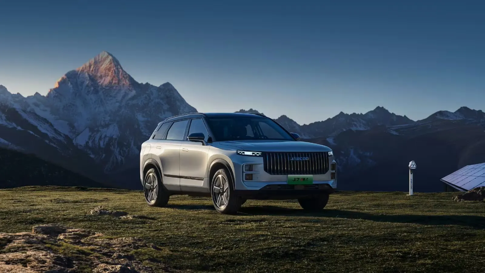 Silver Jaecoo 7 SUV aus dem vorderen Dreiviertelwinkel gesehen, auf Gras geparkt vor einem Hintergrund aus schneebedeckten Bergen.