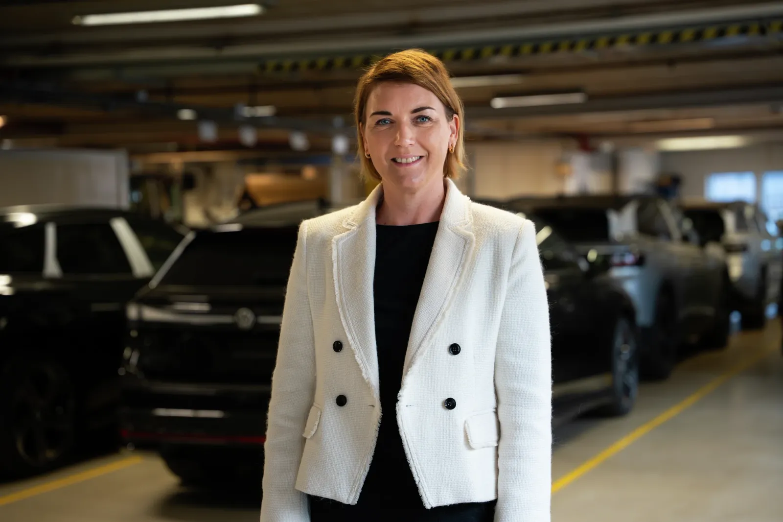 Uma mulher de blazer branco sorri em um estacionamento cercado por carros parados.