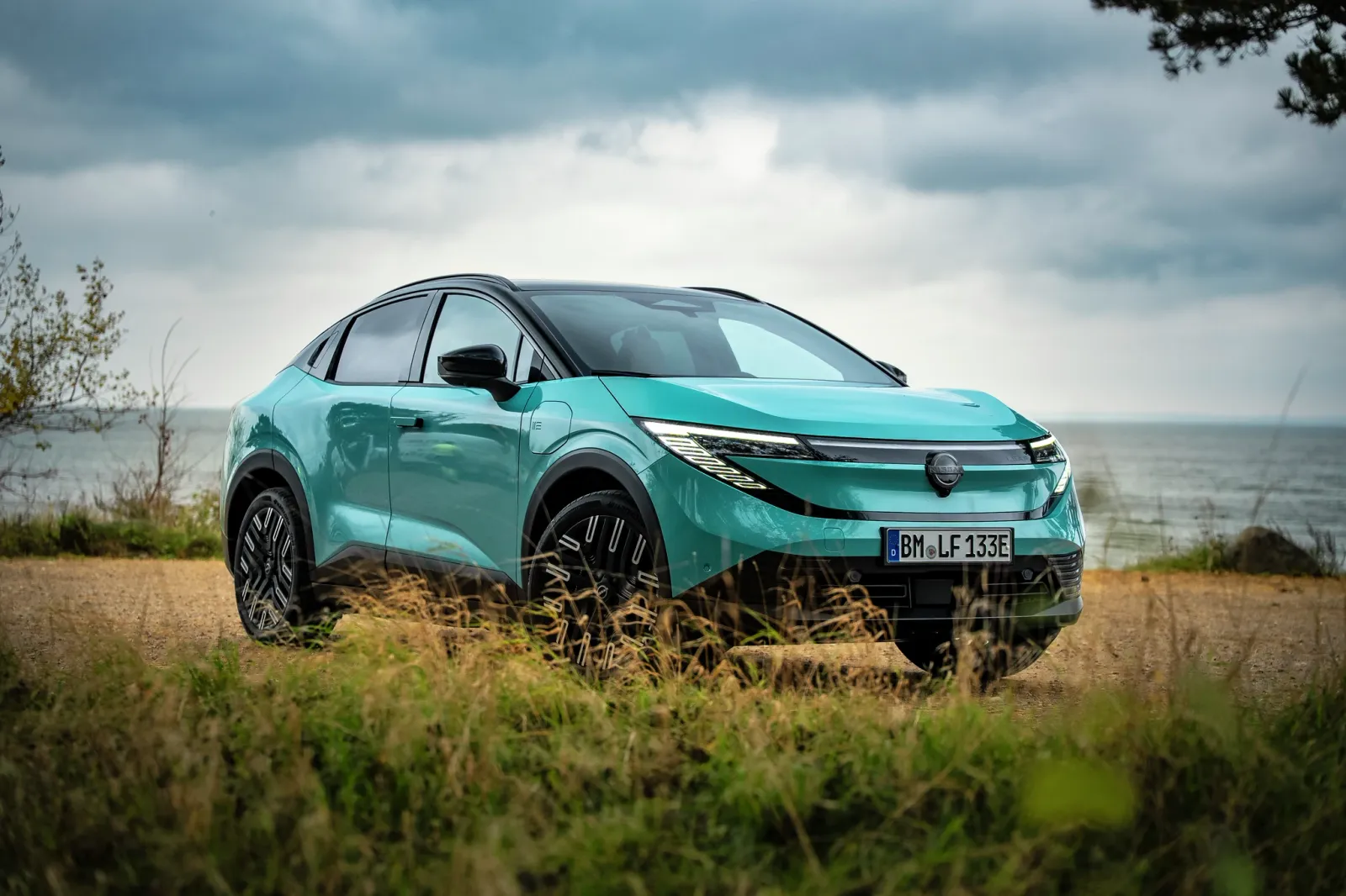 SUV elétrico azul-esverdeado estacionado em uma estrada costeira, com céu e mar ao fundo, demonstrando um design moderno.