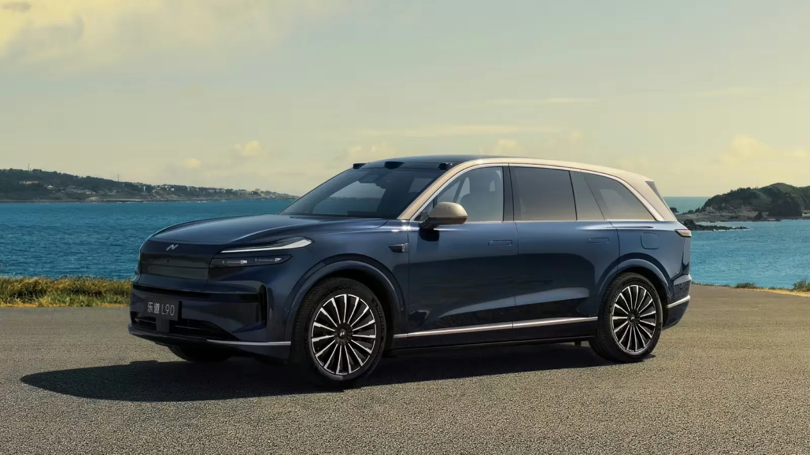 Front three-quarter view of the Onvo L90 with the Starry Gold design package, finished in dark Starry Blue with a gold-toned roof and titanium-colored trim, parked on a seaside road with the ocean and hills in the background.