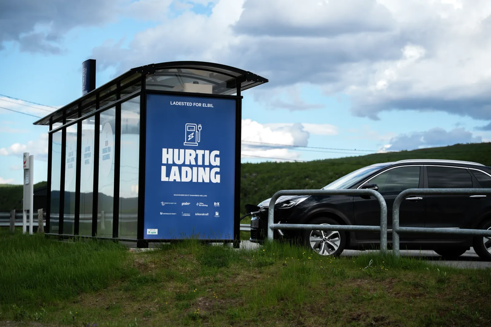 Et busstoppskur med et blått reklameskilt ved siden av en parkert svart bil langs en landlig veikant.