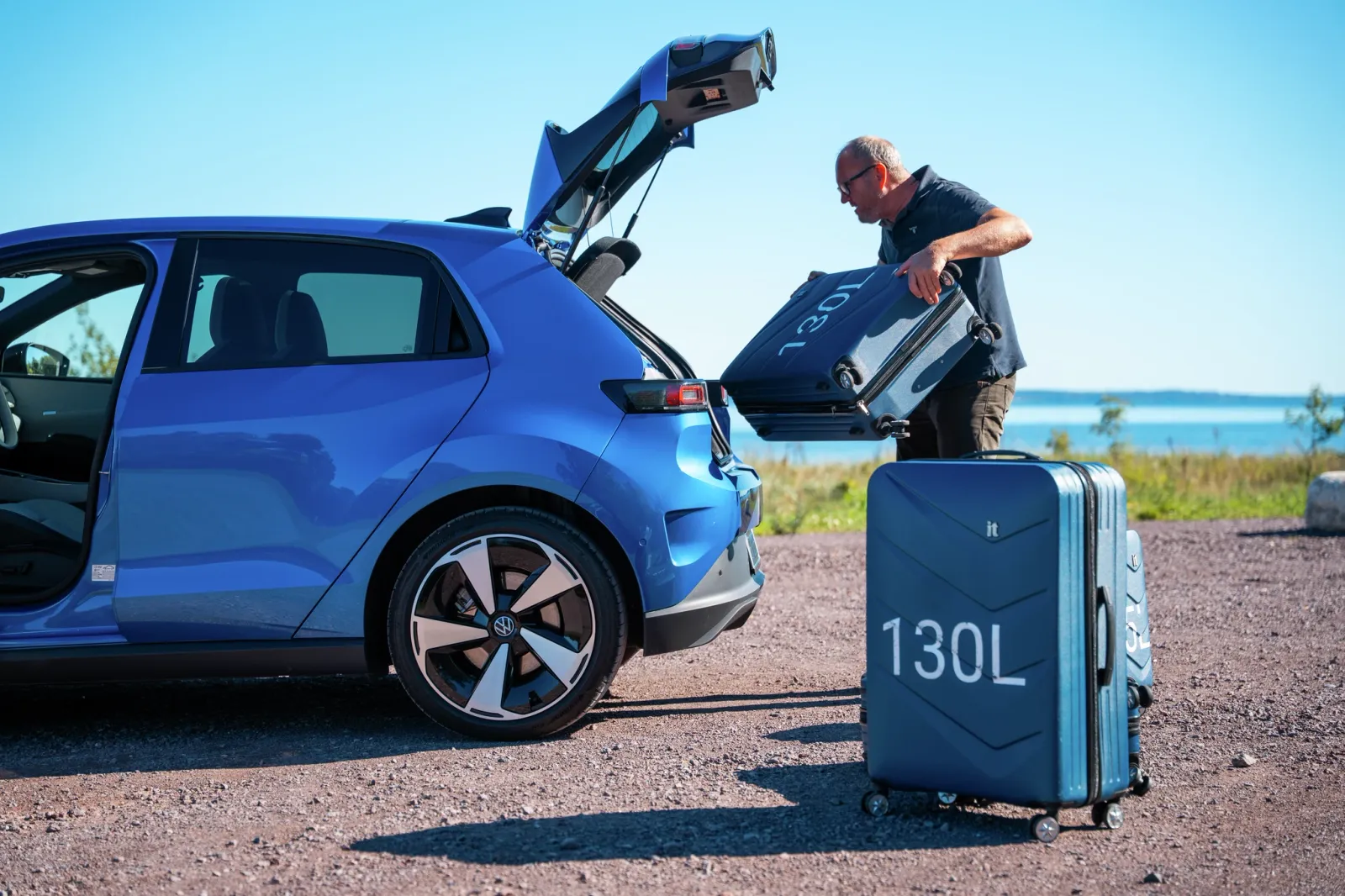 Um homem carrega uma mala de 130 litros no porta-malas de um carro elétrico azul da Volkswagen perto de um lago.