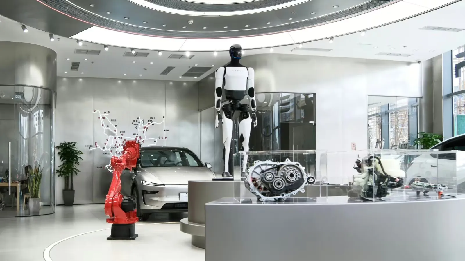 A Tesla Optimus humanoid robot stands on a display platform at a Tesla store in Beijing, next to a red industrial robot arm, a silver Model Y and drivetrain components in glass cases.