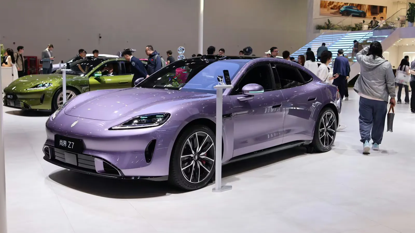 A purple Shangjie Z7 displayed at an auto show, viewed from the front and side, with visitors and a green car in the background.