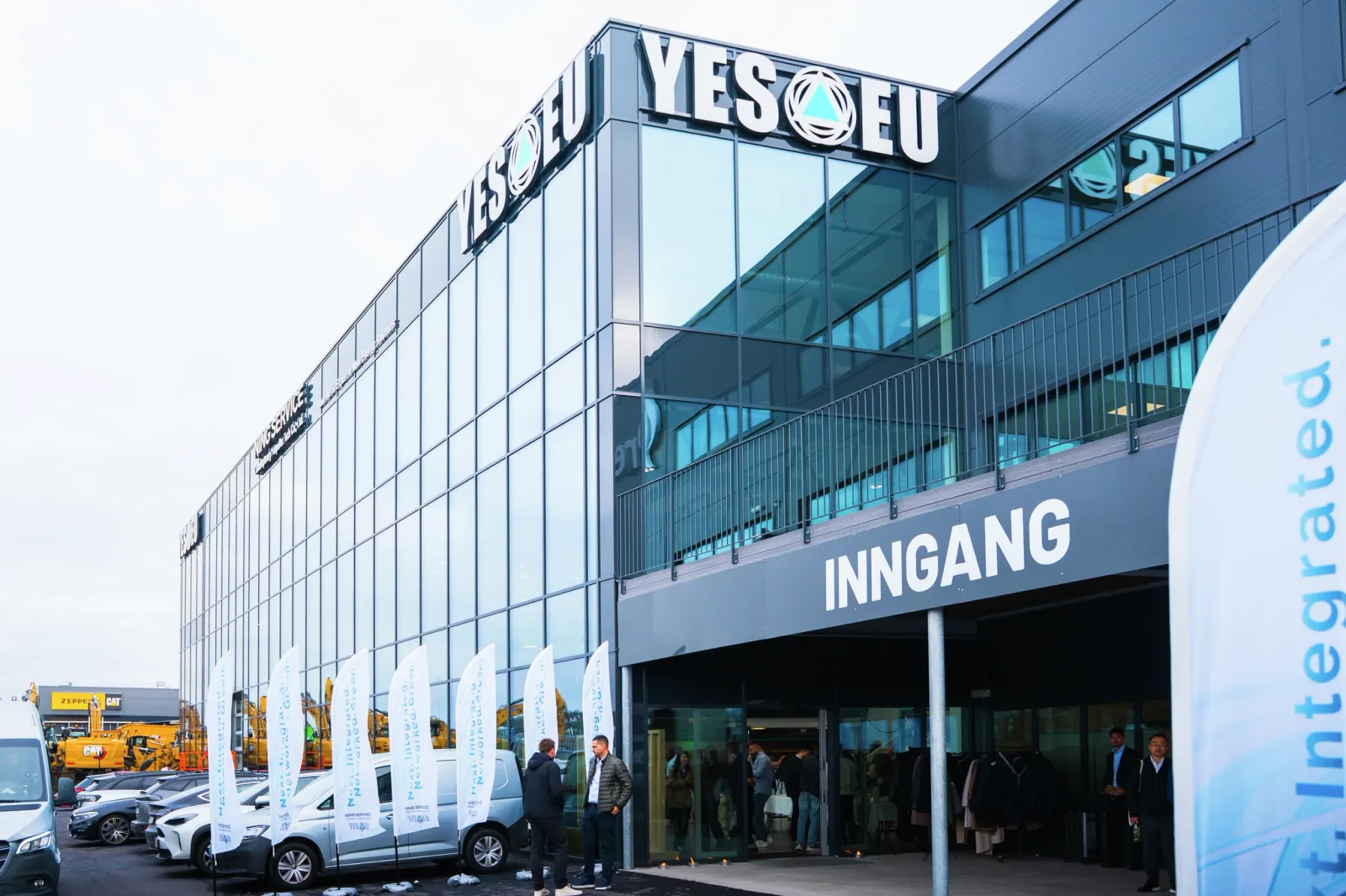 Modern glass building with YES EU mark and ENTRANCE sign, people gathered outside near a flag.